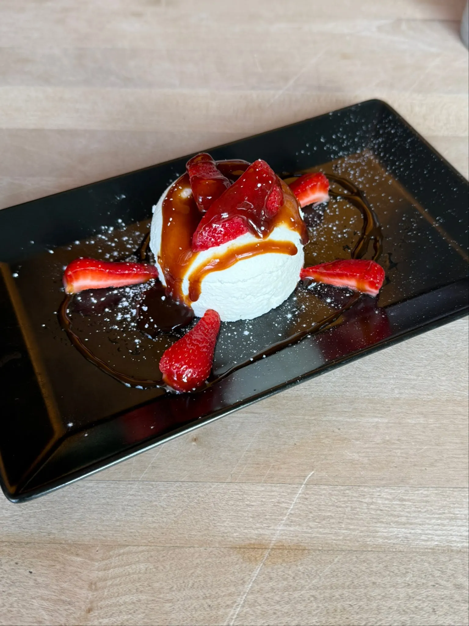 Dessert mit Erdbeeren, Sauce und Puderzucker auf einem schwarzen Teller